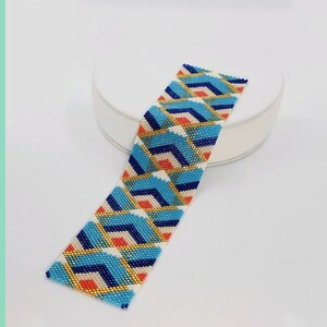 May include: A rectangular beaded bracelet strip featuring a geometric pattern of interlocking chevrons in shades of bright blue, dark blue, cream, and coral, accented with thin diagonal lines of metallic gold beads throughout the design.