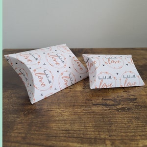 May include: Two white pillow boxes with a repeating pattern of black hearts and the text "beaded with love" in a peachy-pink color. The boxes are on a brown wooden surface.
