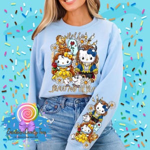 Puede incluir: Una sudadera azul claro con un estampado gr&aacute;fico de Hello Beauty and the Beast que presenta una imagen de dibujos animados de Bella y la Bestia como personajes de Hello Kitty. El gr&aacute;fico tambi&eacute;n incluye una rosa, una corona y un castillo.