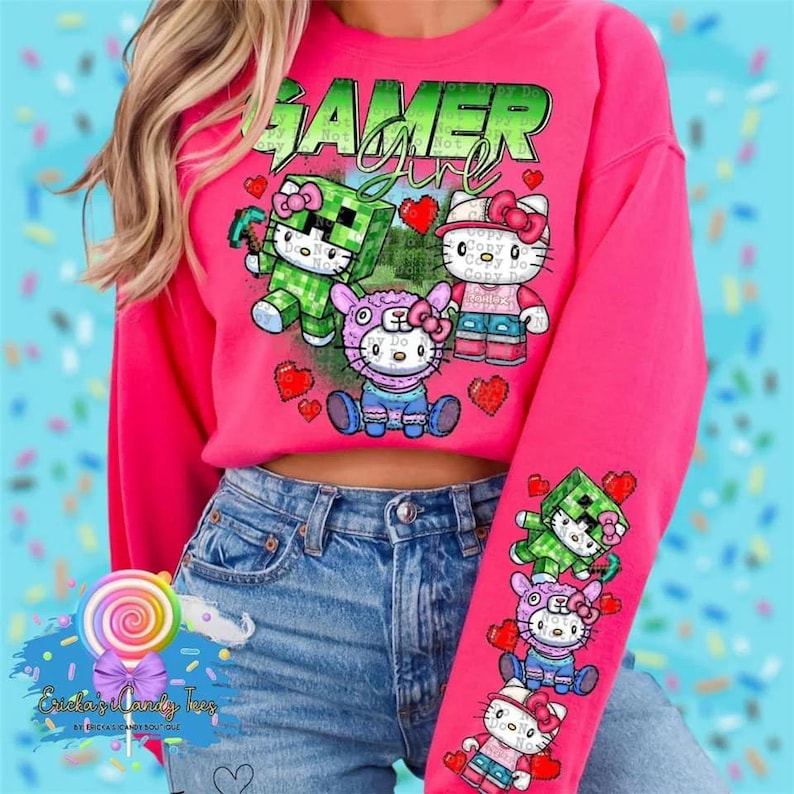 以下が含まれることがあります： ピンクのクロップドスウェットシャツに、ゲームをしている姿のハローキティのキャラクターがプリントされています。スウェットシャツの前には「Gamer Girl」の文字がプリントされています。