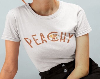 Op de afbeelding: Een wit T-shirt met het woord "PEACHY" bedrukt in bruin met een perzikontwerp in de "Y". Het model draagt ​​een zwarte spijkerbroek en zit op een lichtgrijze ondergrond.