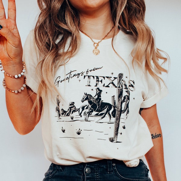 May include: Cream-colored t-shirt with a black graphic of a cowboy on a horse, cacti, and the words "Greetings from TEX". The shirt is paired with dark denim jeans and white shoes. A gold necklace and bracelets complete the look.