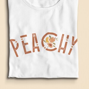 K&ouml;nnte beinhalten: Wei&szlig;es T-Shirt mit dem Wort "Peachy" in Braun bedruckt, wobei ein Pfirsich-Design im "O" enthalten ist.