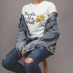 Puede incluir: Una camiseta blanca con el texto "Made in the West" y un gr&aacute;fico de una flor amarilla. La camiseta la lleva puesta una persona que lleva pantalones vaqueros azules y una chaqueta vaquera.