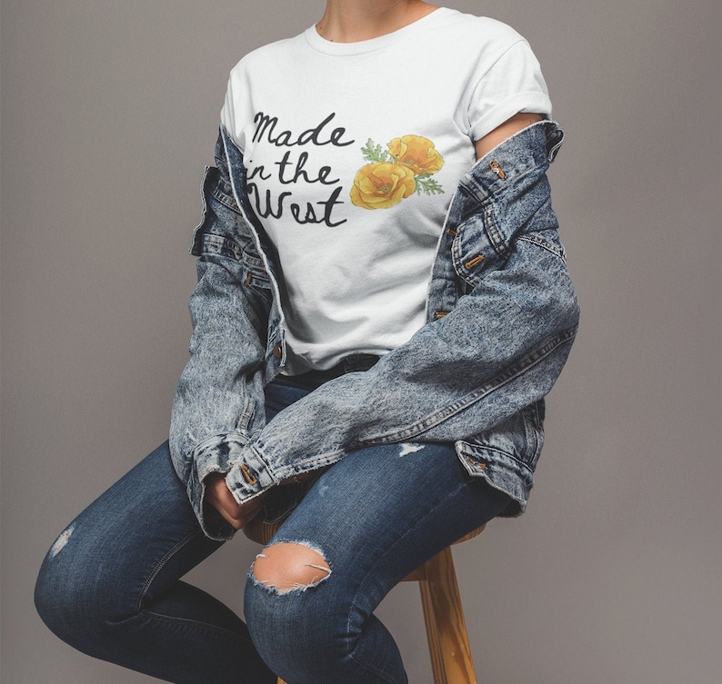 Puede incluir: Una camiseta blanca con el texto "Made in the West" y un gr&aacute;fico de una flor amarilla. La camiseta la lleva puesta una persona que lleva pantalones vaqueros azules y una chaqueta vaquera.