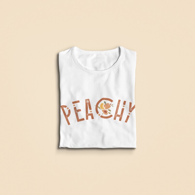 K&ouml;nnte beinhalten: Wei&szlig;es T-Shirt mit dem Wort "Peachy" in Braun bedruckt, wobei ein Pfirsich-Design im "O" enthalten ist.