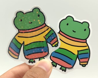 May include: Two die-cut stickers featuring a green frog wearing a rainbow-striped sweater. One sticker has a holographic finish with star-shaped sparkles, while the other has a matte finish. Both stickers include the text LUBLY SHOP.