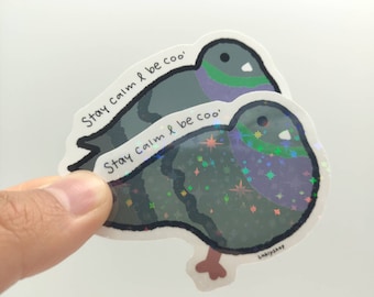 May include: A die-cut sticker featuring a cartoon pigeon with gray, green, and purple plumage. The sticker has a holographic star pattern and the text 'Stay calm & be coo' printed in black. A small brown foot is visible at the bottom edge.
