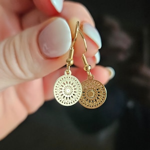 Può includere: Un paio di orecchini dorati con un design circolare delicato. Gli orecchini presentano un motivo a filigrana con un ritaglio circolare al centro.