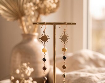 May include: A pair of gold-toned dangle earrings featuring a sunburst charm at the top, followed by a vertical sequence of four multi-colored beads in shades of yellow, black, brown, and grey, ending with a small gold-toned teardrop-shaped metal charm.