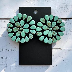 May include: Pair of turquoise-colored plugs, each with a flower-like design. The plugs are made of small, teardrop-shaped stones set on a black base. The stones have a mix of light and dark turquoise hues with brown accents.