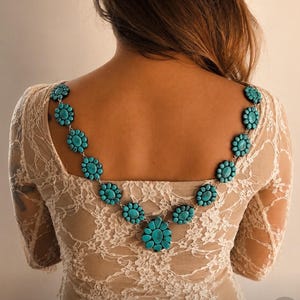 Puede incluir: Un collar que presenta una serie de piedras ovaladas de color turquesa dispuestas en grupos en forma de flor, conectadas por eslabones de metal plateado. La pieza se lleva en la espalda, sobre los hombros de una blusa de encaje beige de manga larga.