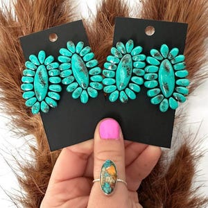 May include: Turquoise-colored stud earrings and a ring with a turquoise stone. The earrings are oval-shaped with a cluster of turquoise stones around a central stone. The ring has a silver band and a similar turquoise stone.