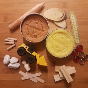 Könnte beinhalten: Eine Sammlung von Kinderspielzeug und Bastelmaterialien, darunter zwei Dosen Knete, ein CAT-Spielzeugbagger, Holzklötze, ein Lineal, Holzdübel und kleine Glasperlen. Die Knetdosen sind mit "HANDMADE WITH LOVE" gestempelt.
