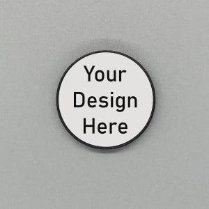 May include: A circular white button with a black edge, featuring the text "Your Design Here" in black. The button is set against a plain grey backdrop.