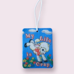 May include: A blue felt car air freshener with a white lamb jumping over a field of flowers. The text "My Life is Crap." is printed on the air freshener.