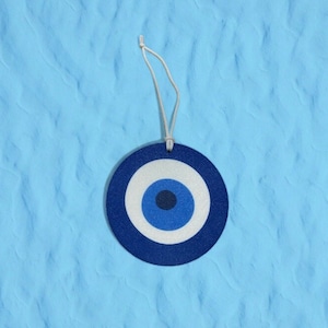 May include: A round, blue and white Evil Eye charm with a string for hanging. The charm features a dark blue outer ring, a white inner ring, and a blue center. The background is a textured light blue.