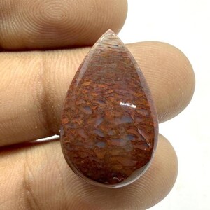 Puede incluir: Una gema pulida en forma de lágrima. La piedra presenta una rica paleta de rojos, marrones y azules. La superficie tiene un patrón texturizado y orgánico. Ideal para joyería o coleccionismo, la gema mide aproximadamente 3 cm de largo.