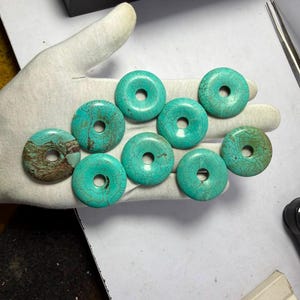 May include: A collection of nine circular turquoise stone beads, each featuring a central hole. The beads display a vibrant teal colour with intricate dark brown veining patterns across their surfaces, varying in intensity and texture.