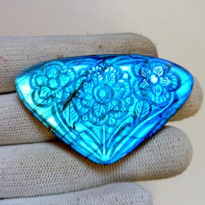 May include: A blue labradorite gemstone with carved floral designs. The gemstone is a vibrant blue with a polished surface. The carved flowers and leaves are visible on the surface. The gemstone is held in a white glove.