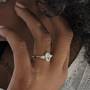 May include: A close-up of a person's hand wearing a silver ring with a large oval-shaped diamond and two smaller diamonds on either side. The ring is on the person's left ring finger.