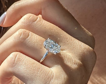 May include: A silver engagement ring with a large, oval-cut diamond. The ring is set on a delicate band and is displayed on a hand with manicured nails. The diamond sparkles in the light, showcasing its facets.