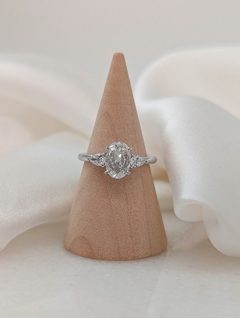 May include: A silver ring with an oval-shaped diamond and two smaller diamonds on either side. The ring is displayed on a wooden ring holder.