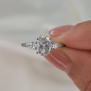 May include: A silver engagement ring with a large oval-shaped diamond and two smaller pear-shaped diamonds on either side.
