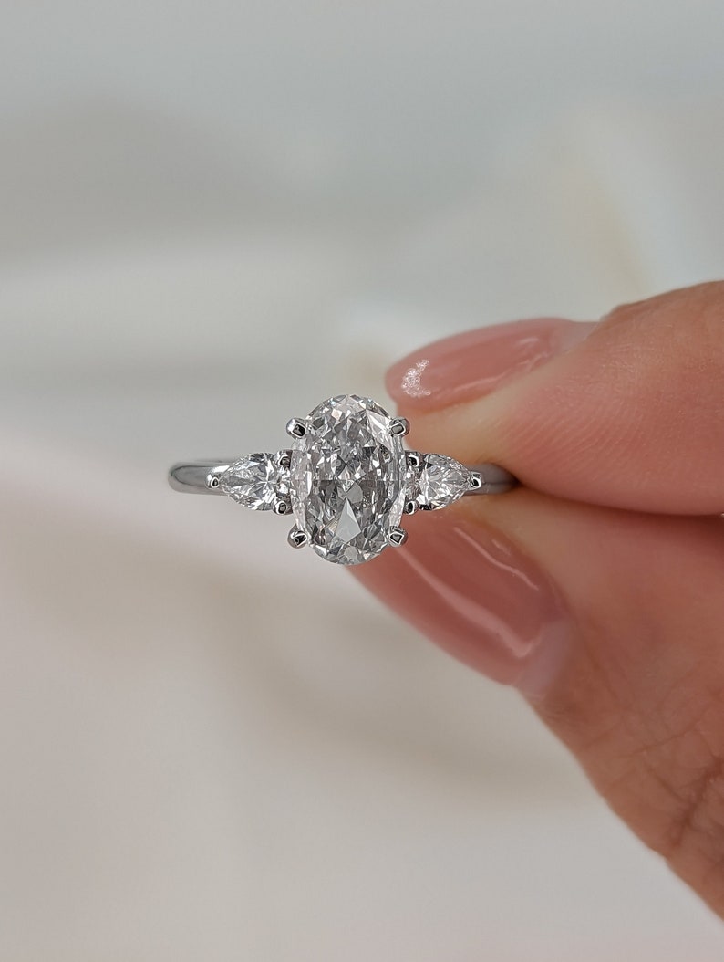May include: A silver engagement ring with a large oval-shaped diamond and two smaller pear-shaped diamonds on either side.