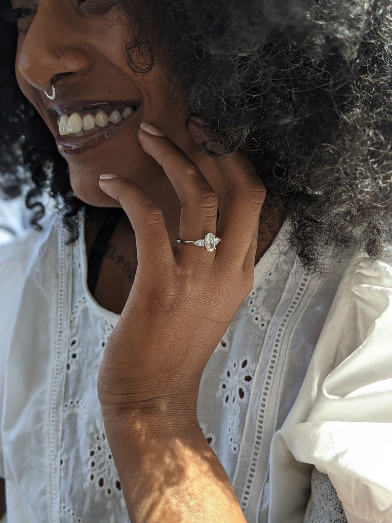 May include: A close-up of a person's hand wearing a silver ring with a large oval-shaped diamond and two smaller diamonds on either side. The ring is on the person's left ring finger.