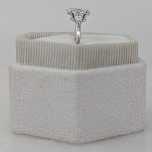 May include: A silver ring with a large, clear, emerald-cut diamond surrounded by smaller diamonds. The ring is displayed in a white velvet ring box.