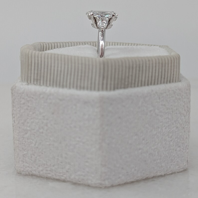 May include: A silver ring with a large, clear, emerald-cut diamond surrounded by smaller diamonds. The ring is displayed in a white velvet ring box.