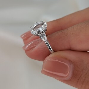 May include: A silver engagement ring with an oval-shaped center stone and two smaller stones on either side. The ring is set on a thin band.