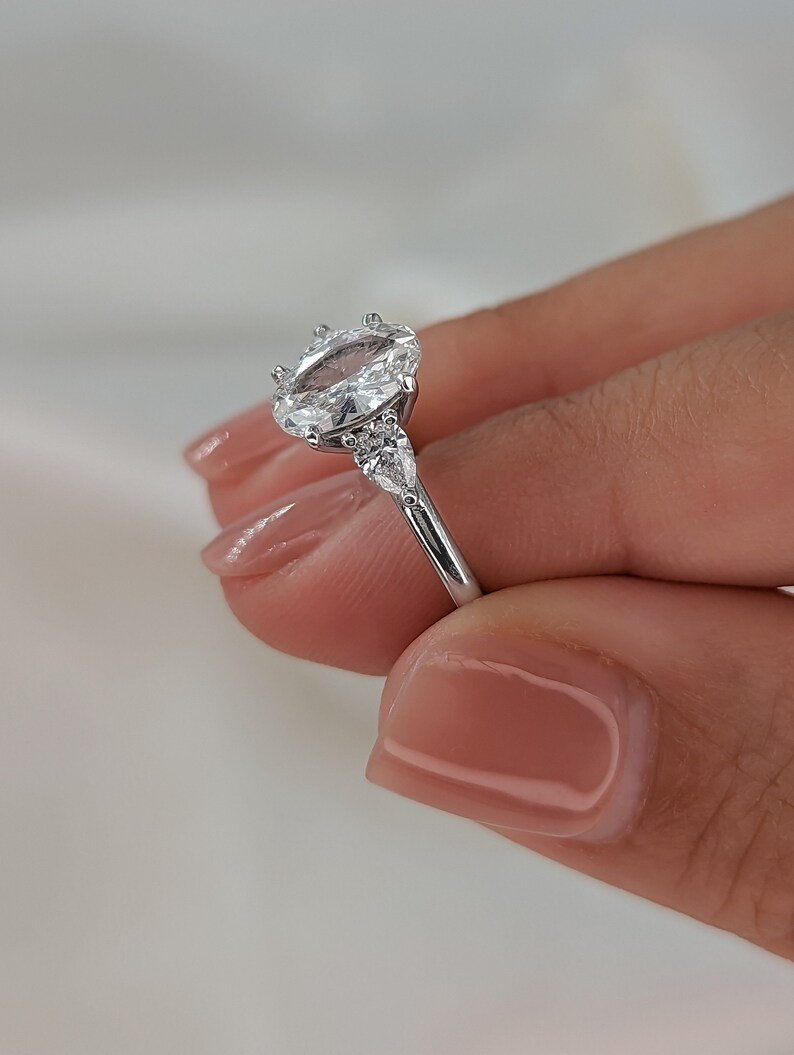 May include: A silver engagement ring with an oval-shaped center stone and two smaller stones on either side. The ring is set on a thin band.