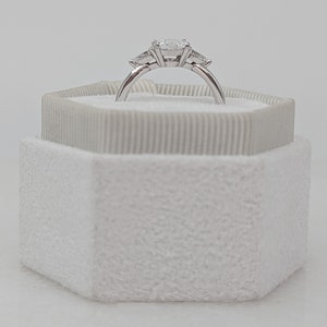 May include: A white ring box with a white velvet interior. A silver ring with three diamonds is displayed inside the box.