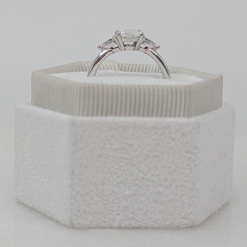 May include: A white ring box with a white velvet interior. A silver ring with three diamonds is displayed inside the box.