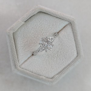 May include: A silver ring with an oval-shaped center diamond and two smaller pear-shaped diamonds on either side. The ring is in a white ring box.
