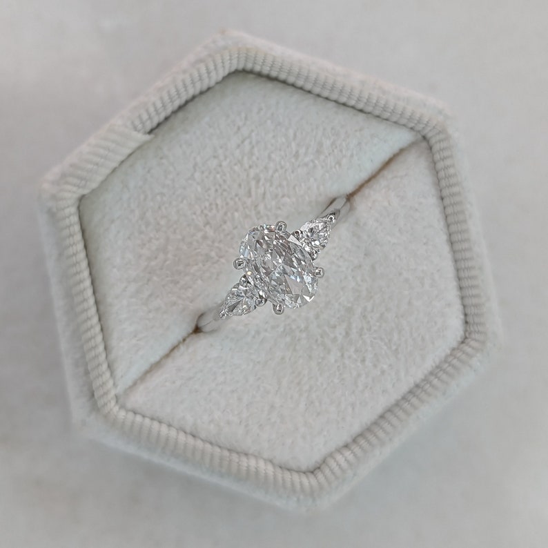 May include: A silver ring with an oval-shaped center diamond and two smaller pear-shaped diamonds on either side. The ring is in a white ring box.