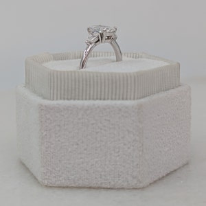 May include: A pear-shaped diamond engagement ring with two smaller diamonds on the band, set in a white gold setting. The ring is displayed in a white velvet ring box.