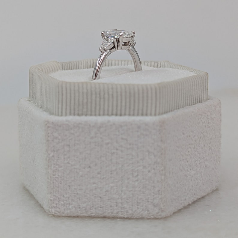 May include: A pear-shaped diamond engagement ring with two smaller diamonds on the band, set in a white gold setting. The ring is displayed in a white velvet ring box.