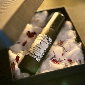 Puede incluir: Un pequeño frasco de vidrio verde oscuro con una etiqueta blanca que dice "Dream Worker Scented Perfume Oil" en una fuente decorativa. El frasco está en una caja negra con algodón blanco y pétalos de flores rojas y azules.