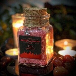 May include: A glass jar filled with pink salt, labeled "Midnight Imperium Fire Ritual Salt". The jar has a cork lid and a silver charm attached to it.