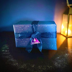 May include: A wrapped gift box in shades of blue, adorned with a dark blue ribbon and a tag that reads "Yule Blessings." The wrapping paper features a pattern of small white stars. The overall aesthetic suggests a festive or holiday theme.