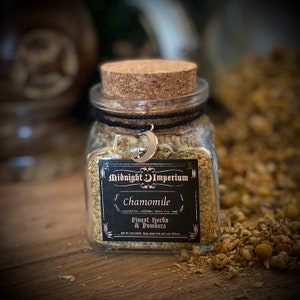 Puede incluir: Un frasco de vidrio con tapa de corcho lleno de flores de manzanilla secas. El frasco tiene una etiqueta negra con el texto "Midnight Imperium Chamomile Finest Herbs & Powders" y un s&iacute;mbolo de luna creciente.