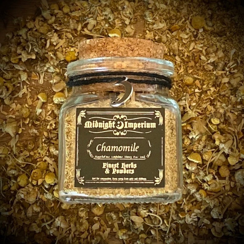 Puede incluir: Un frasco de vidrio con tapa de corcho lleno de flores de manzanilla secas. El frasco tiene una etiqueta negra con el texto "Midnight Imperium" y "Chamomile" en letras blancas. La etiqueta tambi&eacute;n dice "Finest Herbs & Powders".