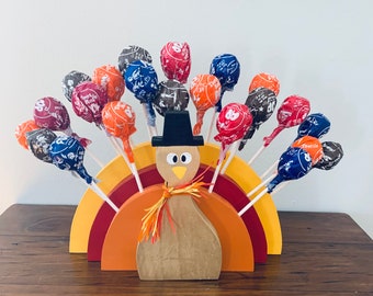 May include: A wooden turkey figurine with a colorful lollipop tail. The turkey is brown and has a black hat. The lollipops are red, blue, orange, and yellow.