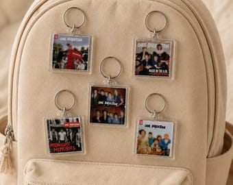 以下が含まれることがあります： One Directionのアルバムジャケットがデザインされた5つの正方形の透明アクリルキーホルダー。Midnight MemoriesやMade in the A.M.などが含まれ、ベージュのバックパックに取り付けられています。各キーホルダーには銀色の金属リングが付いており、バンドの写真が印刷されています。