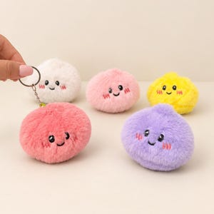May include: A collection of five round, plush keychains in white, pink, yellow, coral, and purple. Each fuzzy ball features embroidered black eyes, a small smile, and pink stitched cheeks. One coral keychain is attached to a silver metal ring.