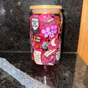 May include: A glass water bottle with a pink background and various illustrations, including a shield with the text "Basgiath War College", a feather, a dagger, a potion bottle, a book, a crescent moon, and the text "I will not die today".