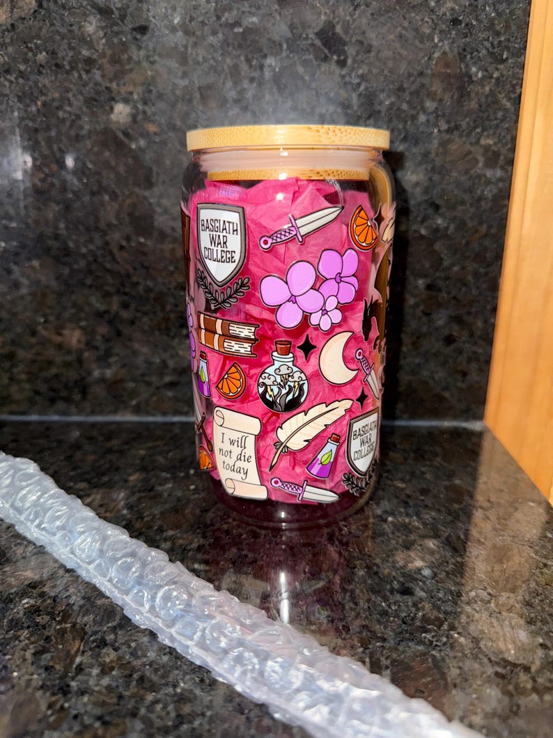 May include: A glass water bottle with a pink background and various illustrations, including a shield with the text "Basgiath War College", a feather, a dagger, a potion bottle, a book, a crescent moon, and the text "I will not die today".